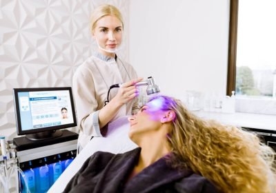 Beautician doctor doing light rejuvenation for pretty young woman at beauty salon clinic. Aesthetic facial light skin treatment. Hardware cosmetology.